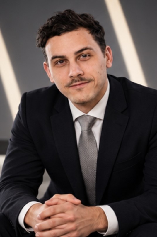 Maxime seated, hands clasped, smiling